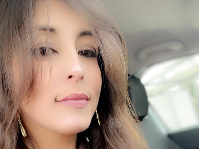 Lolita123456 has a cute face with wavy brown hair, light skin, and pink lips, wearing gold earrings, sitting in a car, looking down with a subtle smile.
