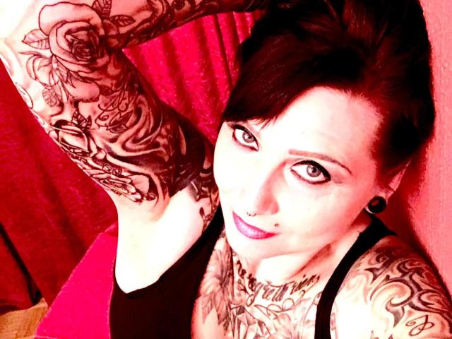 StrengeTante-hot with tattoos, dark hair, purple lipstick, nose ring, and black ear plugs, raising arm, wearing black tank top, showing tattoos on arm and chest.