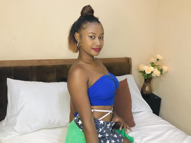 JulianaSabi has brown skin, red lipstick, and a bun, wearing a blue strapless top and star-patterned skirt, sitting on a bed with white pillows and brown headboard.