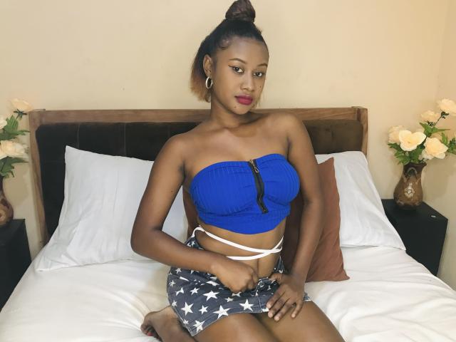 JulianaSabi, with dark skin, wearing a blue strapless top and star-patterned shorts, sitting on a bed, showing her small tits and flat ass, with her hand on her crotch.