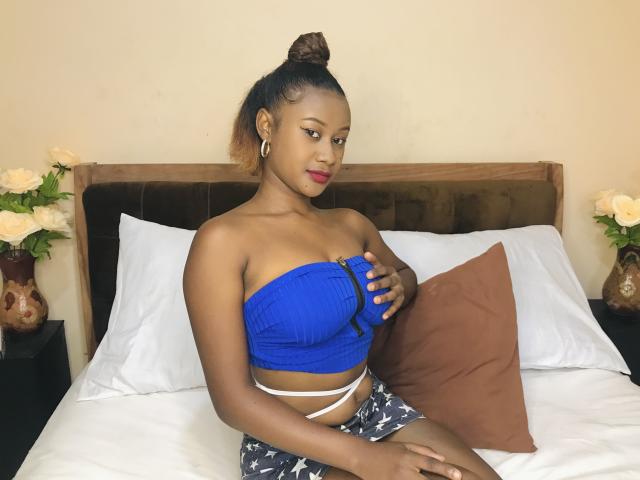 JulianaSabi with dark brown skin, small tits, blue strapless top, black patterned shorts, brown bun, red lipstick, holding brown pillow on white bed with brown headboard.