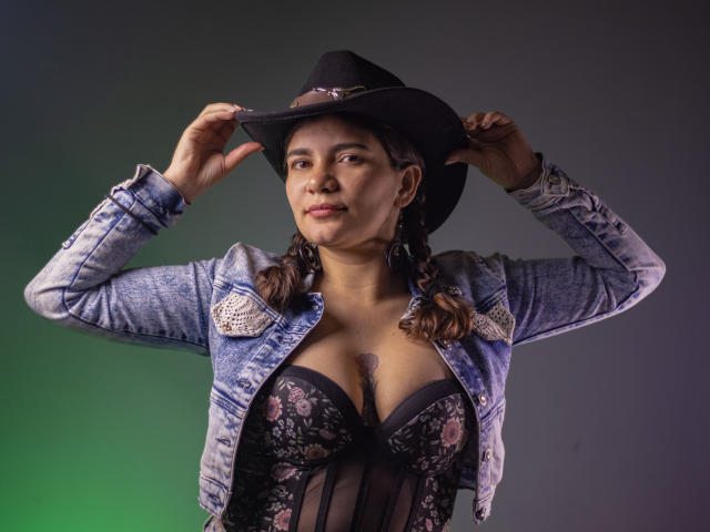 BulmaHot wears a black cowboy hat, blue denim jacket, and black floral lingerie with visible boobs, adjusting the hat with a confident look, brown braids, green and gray background.