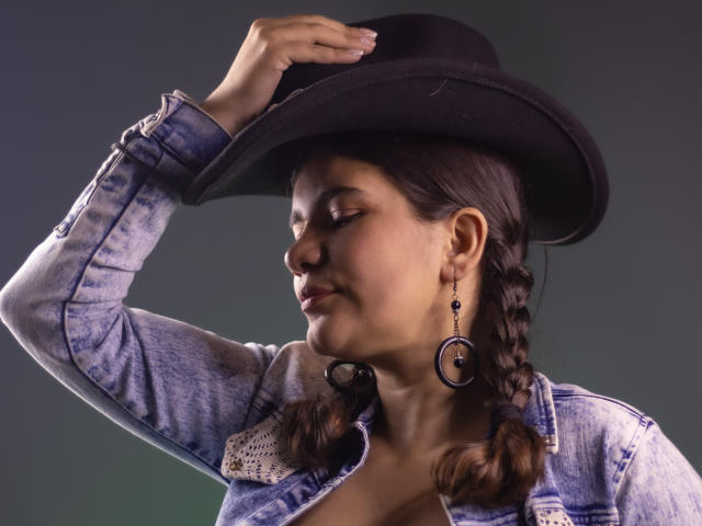 Bulma, con el cabello castaño trenzado, usando un sombrero de vaquero negro, una chaqueta vaquera azul y aros morados, tocándose el sombrero con la mano derecha, con los ojos cerrados..