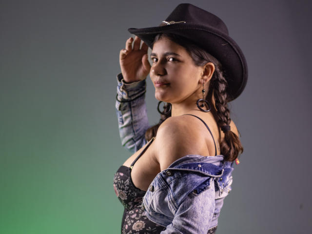 BulmaHot with braided hair, black cowboy hat, denim jacket, and floral lace-up top, showing off her boobs, looking over shoulder, green-gray gradient background.