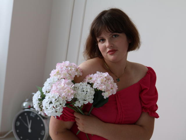 StaceyLex com flores rosa e brancas, segurando-as contra o peito, vestindo uma blusa vermelha com um ombro de fora, cabelo castanho, sorriso discreto, colar verde, relógio ao fundo..
