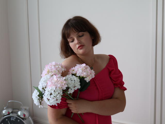 StaceyLex with short brown hair, wearing a red dress, holding a bouquet of pink and white hydrangeas, looking down, standing in a white room, near an alarm clock.