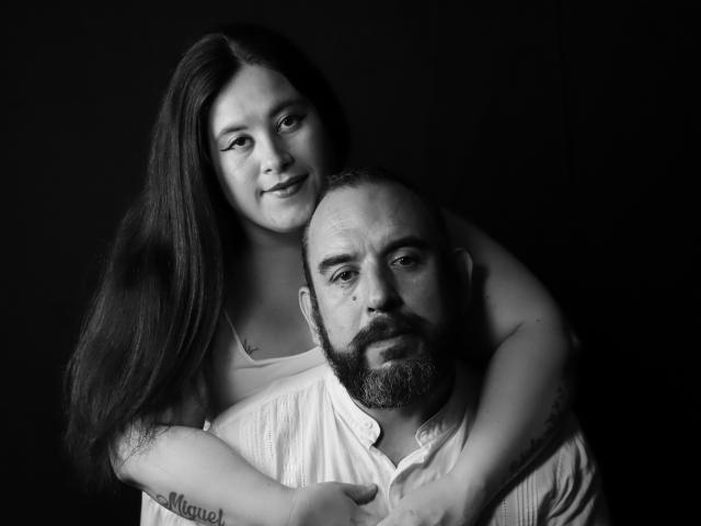 SarahMat met lang donker haar, armen om een kale, baardige man in een wit shirt, die naar de camera kijkt. Haar tatoeage "Manel" is zichtbaar op haar onderarm. Zwart-wit, intieme pose..