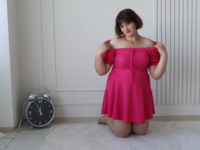 StaceyLex ajoelhada em um tapete branco, vestindo um vestido rosa com um ombro descoberto, segurando as alças do vestido, com cabelo castanho curto, um colar pequeno e um despertador de prata à esquerda..