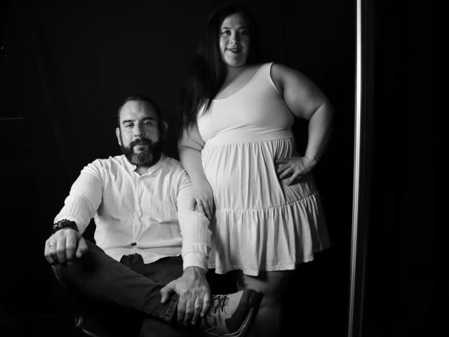 SarahMat with big tits stands, hands on hips, wearing a white dress. Bearded dude with a beard sits, legs crossed, wearing a white shirt and dark pants. Black and white.