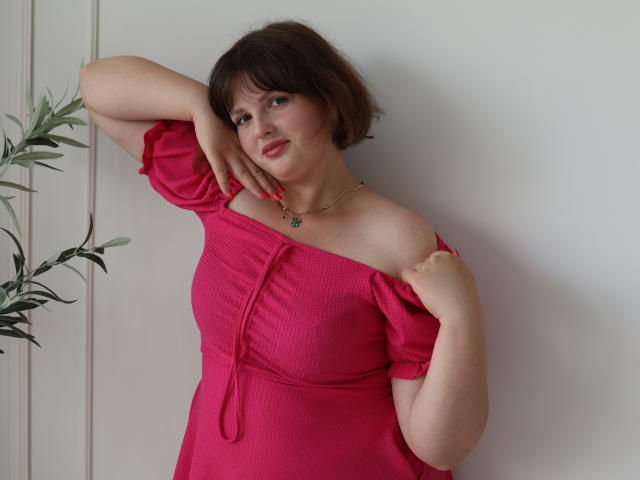 StaceyLex with short brown hair, pink off-shoulder dress, holding one arm up, showing her tits, smiling, green leafy plant on left, white wall, gold necklace.