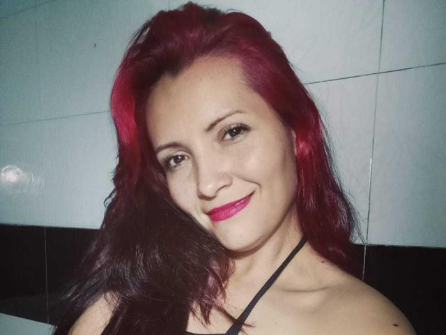 Natnys has red and black hair, smiling with pink lipstick, wearing a black halter top, showing her tits, and has light skin and brown eyes.