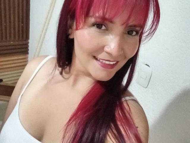 Natnys with red and black hair, smiling, wearing a white tank top, showing her tits, nose ring, and pink lipstick. She has light skin and brown eyes.