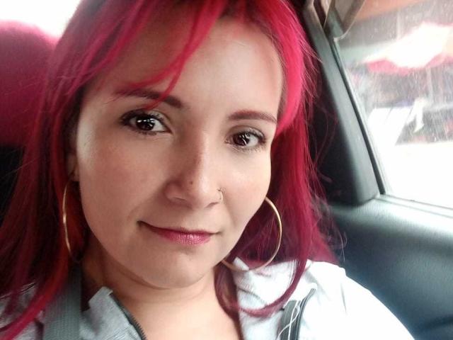 Natnys with red hair, small nose ring, and hoop earrings, smiling in a car, wearing a gray zip-up jacket, brown eyes, slight smile, pinkish lips.