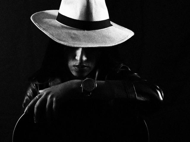 MichaelaMistress with dark hair, wearing a white cowboy hat and black leather jacket, arms crossed, holding a guitar, wristwatch on her right hand, in a black-and-white shot.