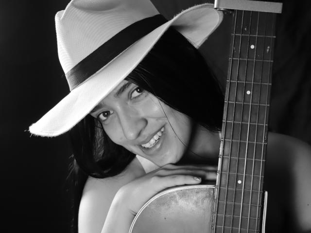 MichaelaMistress with long black hair, wearing a white hat with a black band, smiling, holding a guitar, showing her tits and ass, in a black-and-white photo.