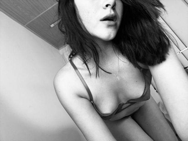 SpencerLe with dark hair, wearing a grey bra, showing her tits, slightly open mouth, necklace with a star pendant, leaning forward, black and white photo.