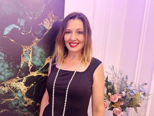 NikaStraitlive with blonde ombre hair, black sleeveless dress, white pearl necklace, red lipstick, smiling, standing next to floral arrangement, green and gold patterned wall.