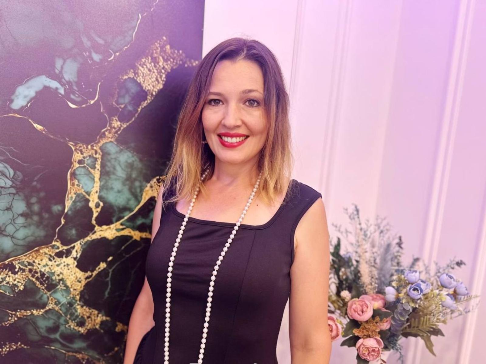 NikaStraitlive with blonde ombre hair, black sleeveless dress, white pearl necklace, red lipstick, smiling, standing next to floral arrangement, green and gold patterned wall.