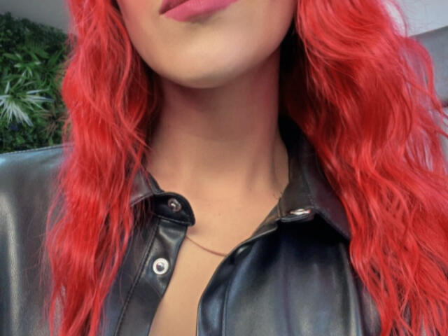 AlissaBrown with bright red, wavy hair, wearing a black leather jacket, showing a bit of her chest, and pink lips. Her neck and upper chest are visible.