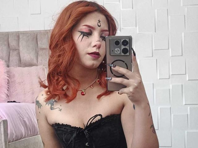 EmmaThompsonn with red hair, black lace top, tattoos, goth makeup, holding phone, taking selfie, wearing red necklace, showing her tits, in mirror selfie.