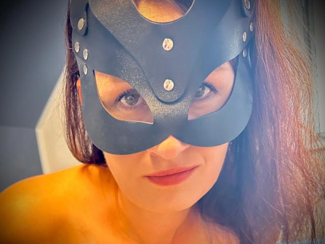 Jacky8890-hot with a black leather mask, brown hair, and a slight smile, showing her eyes and lips, wearing an orange top, in a well-lit, warm-toned photo.