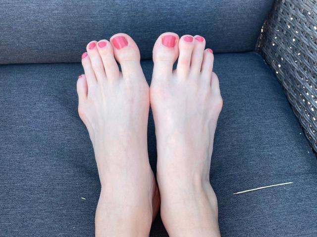 Jacky8890-hot's pale feet with pink toenails, toes pointed slightly upward, resting on a gray wicker couch. Visible tan line on right ankle.