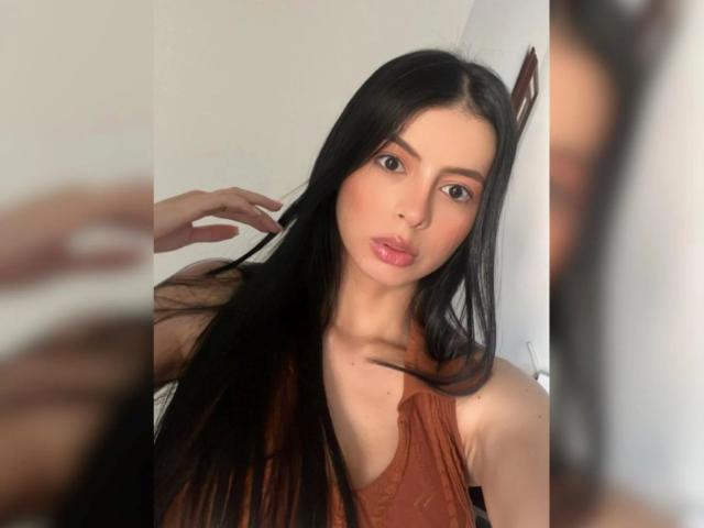 IsabellaJames with long black hair, brown eyes, and pink lips, wearing a brown sleeveless top, touching her hair. Blurred reflection in the mirror.