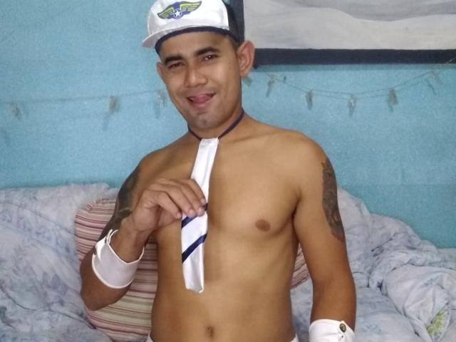 Jeciel, shirtless, wearing a white cap with a blue and green emblem, holding a white and blue striped necktie, tattoos on right arm, wristbands, smiling, tongue out.
