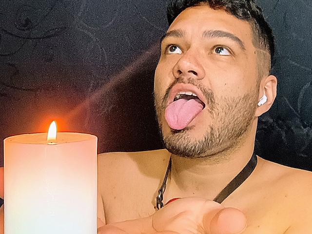 StevenHills with a beard, short curly hair, and white earbuds, sticking out his tongue, holding a lit candle, and wearing a black leather necklace.