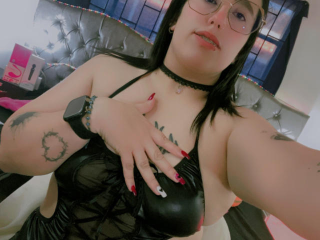 NatalieHarris with black leather dress, glasses, choker, red nails, tattoos, hand on tits, nipple visible, black hair, red lipstick, wrist bracelet, looking at camera, bedroom.