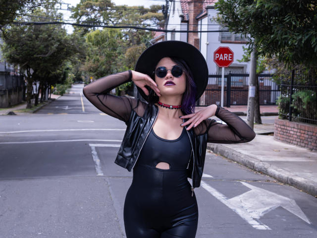 CassieJones with purple hair, black leather jacket, sheer sleeves, black hat, round sunglasses, black cutout top, and black pants, standing on a street with a stop sign.