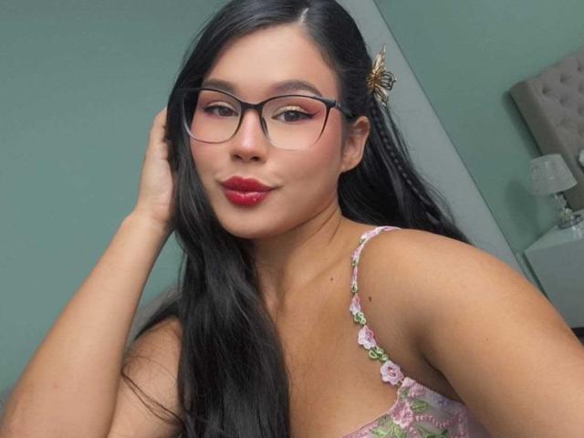CarolineWill with long black hair, wearing glasses, pink lace top, and red lipstick, has a butterfly hairpin, smiling slightly, hand in hair, green wall in background.