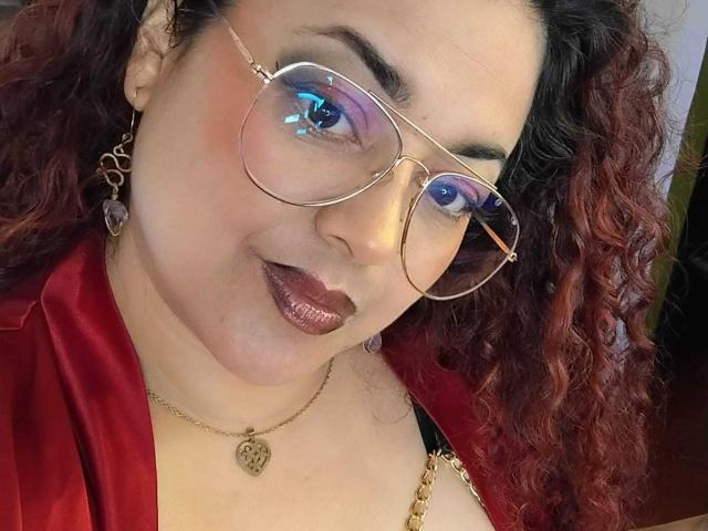 Lalitzfet met krullend rood haar, een bril die blauw licht reflecteert, een rode satijnen robe, donkere lipstick, een gouden ketting en paarse oorbellen, lachend naar de camera. Borsten gedeeltelijk zichtbaar, vanaf de borst naar beneden..