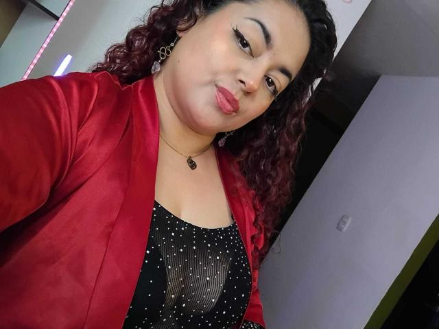 Lalitzfet with curly red hair, red jacket, black sparkly top, puffy lips, necklace, and earrings, showing her tits slightly through her top, taking a selfie.