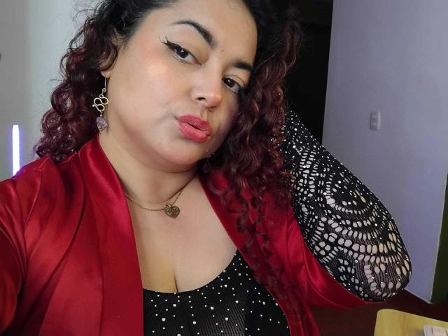Lalitzfet a les cheveux roux bouclés, elle porte une veste en satin rouge par-dessus un haut noir à pois avec des manches en dentelle, elle a les lèvres pincées avec du rouge à lèvres rose et porte un collier en forme de cœur..