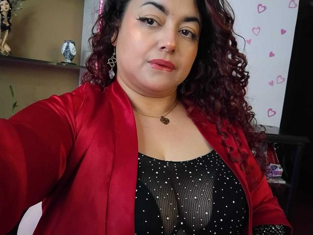 Lalitzfet, con i capelli rossi ricci, una giacca rossa, una maglietta nera con pois bianchi, orecchini a forma di cuore e rossetto rosso, guarda la fotocamera con un leggero sorriso, mostrando il suo seno di medie dimensioni..