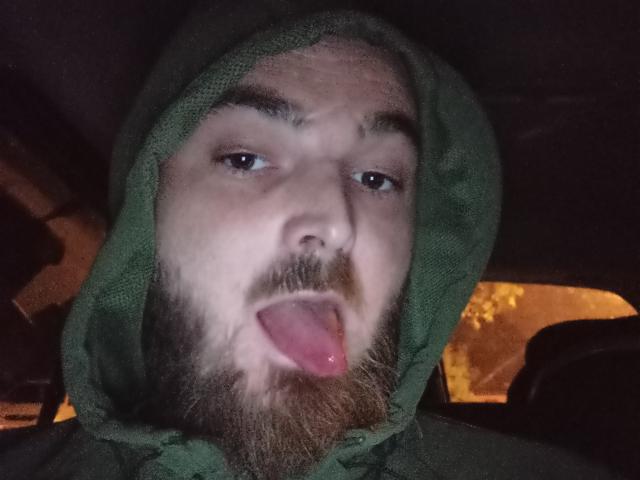 Francais69 mit einer grünen Kapuze, einem braunen Bart und herausgesteckter Zunge, sieht albern in einem dunklen Auto bei Nacht aus. Seine Augen sind halb geöffnet und sein Gesicht ist leicht beleuchtet..