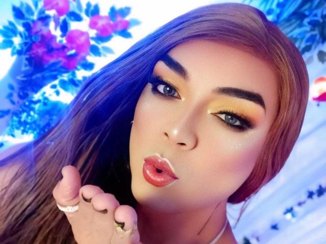 KarolElegance has pouty pink lips, bold orange eyeshadow, and long brown hair. She's making a kissy face with a ringed hand, her blue eyes looking at the camera.