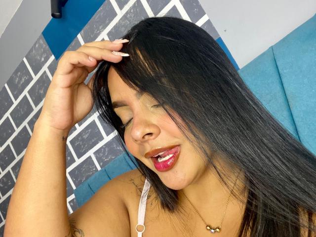 AmarantaCarey has long black hair, tan skin, and pink lipstick, tongue out, hand in hair, wearing a white strap, gold necklace, and blue cushioned background.