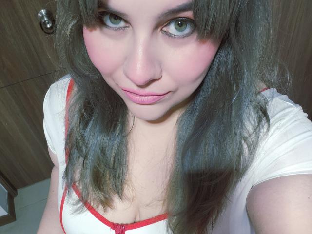 Akkanee, avec ses yeux verts, ses lèvres roses et ses cheveux gris, porte un haut blanc avec des détails rouges, laissant entrevoir sa poitrine, et sourit à l'appareil photo. Ses seins sont légèrement visibles..