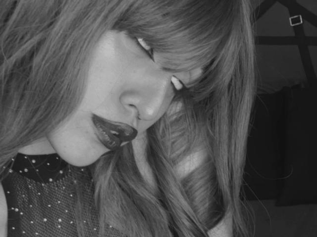 KaylinKrof with long, straight hair, dark lipstick, closed eyes, and a mesh top with small studs. Her face is tilted down, showing her nose and slightly parted lips.