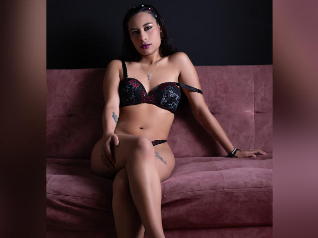 Kate Donovan, con cabello oscuro, lápiz labial morado, un sujetador de encaje negro y rojo, ropa interior negra, tatuajes en el brazo y el muslo, sentada en un sofá rosa, con un top negro de tirantes y las piernas cruzadas..