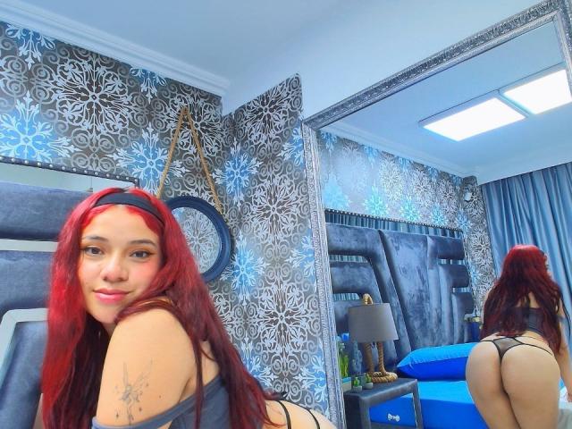 MiaLeroy with red hair, black thong, and tattoo, sitting on bed, showing her ass in mirror reflection, smiling, blue and grey room, bright ceiling light.