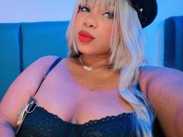 LizzyFoxy with blonde hair, red lipstick, black lace bra, cleavage, necklace, black cap, blue background, and a confident smile.