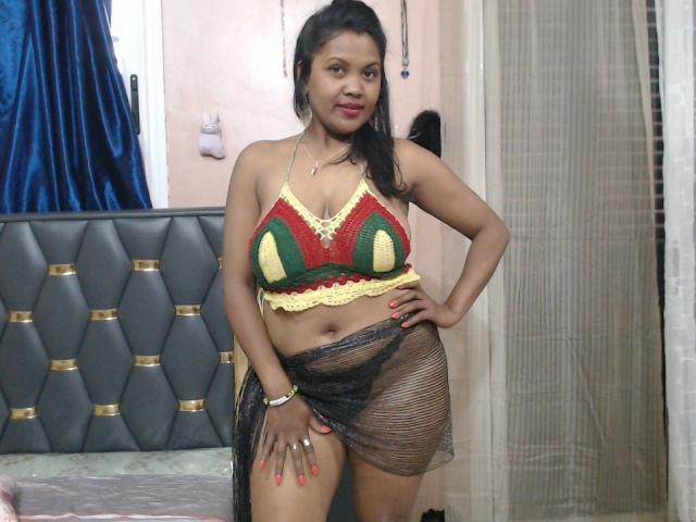 FelyLove with medium brown skin and black hair, wearing a green, red, and yellow crochet top, black mesh skirt, and red nails, standing confidently in a bedroom.