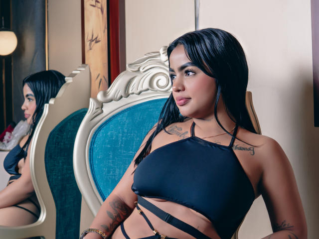 GiaAttum, com cabelo preto, vestindo uma blusa preta com alças finas, mostrando seios pequenos e tatuagens, sentada em uma cadeira azul estofada, olhando para um espelho. Seu reflexo é visível..