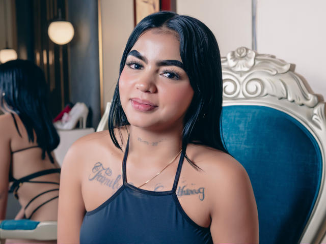GiaAttum ha i capelli neri, le sopracciglia scure e dei tatuaggi sopra il seno con le scritte "Per te" e "Per sempre". Indossa un top nero con spalline e sta seduta su una sedia blu..