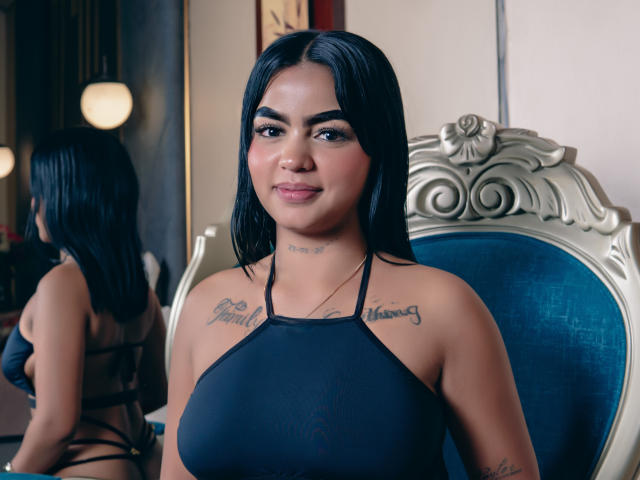 GiaAttum, con cabello negro, busto medio, tatuajes en el pecho, vestida con un top de tirantes negro, sentada en una silla de terciopelo azul, un espejo refleja su espalda. Sonrisa suave, segura de sí misma..