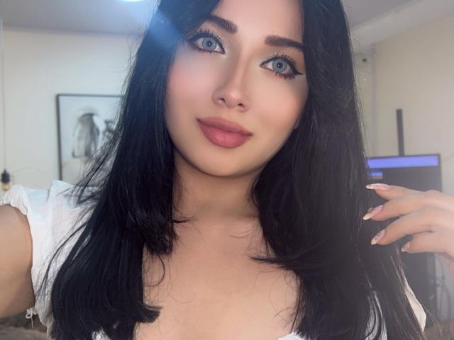 LillyTaylor has blue eyes, black hair, and is wearing pink lipstick. She's smiling with a hand in her hair, showing off her long, manicured nails. She's wearing a white top.