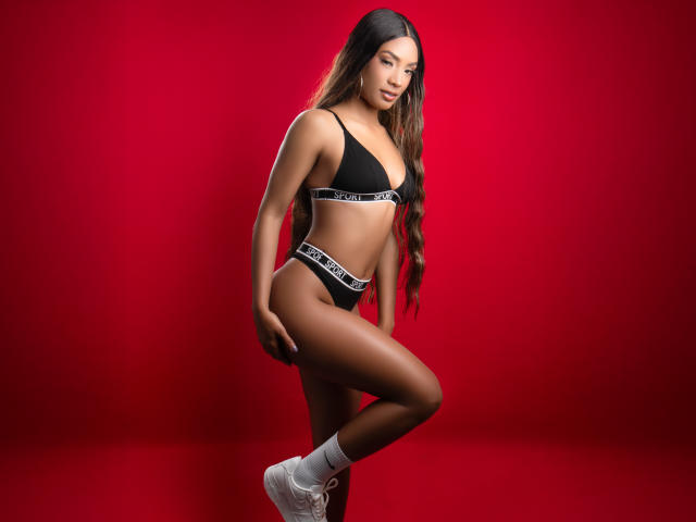 NatashaLewis with long brown hair, wearing black bra and panties, white socks, and white sneakers, standing on one leg, showing her ass and small tits against a red background.